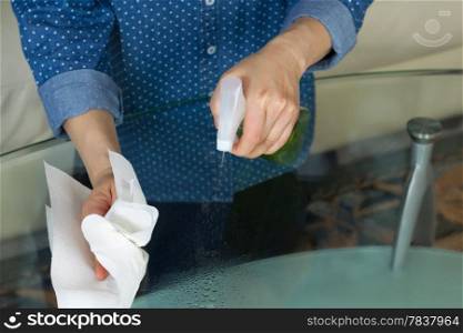 Horizontal photo female hands spraying cleaning solution, from spray bottle, onto dirty glass table with paper towels and sofa in background