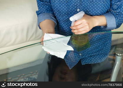 Horizontal photo female hands wiping underneath glass table with spraying cleaning solution bottle and paper towels in hand with sofa in background