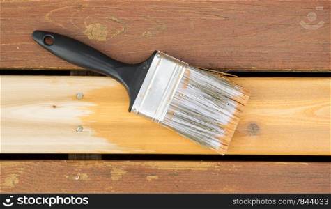 Horizontal photo of a large nylon paint brush on top of new wood board showing cedar stain color next to faded wood on outdoor deck