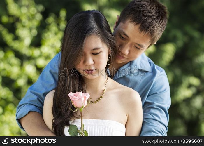 Horizontal photo of a young adult woman holding a pink rose with her lover behind on a blurred green background