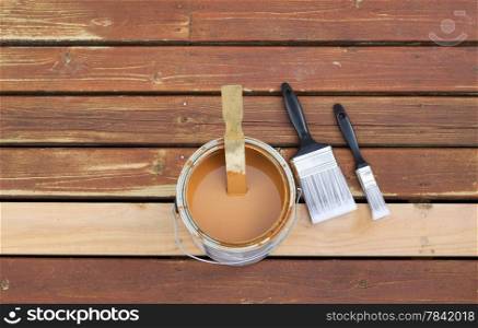 Horizontal photo of an open can of wood stain with two paint brushes lying on a single new cedar wooden board next to fading wood on outdoor deck