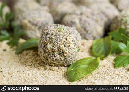 Horizontal photo of bread coated raw meatball on bread crumbs, basil, parsley and finished meatballs in background