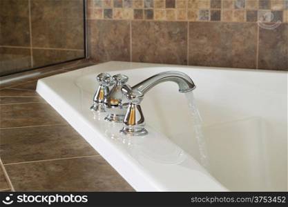Horizontal photo of chrome faucet running water into soaking tub in master bathroom with partial shower glass in background