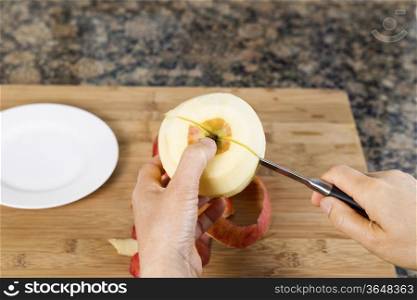 Horizontal photo of female hands cutting freshly peeled apple using a paring knife with bamboo cutting board, white dish and stone counter top in background