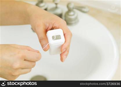 Horizontal photo of female hands pulling dental floss out of container in bathroom with sink and counter top in background