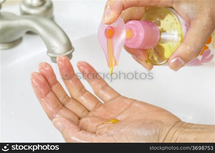 Horizontal photo of female hands putting liquid wash soap, from bottle, into hand with bathroom sink and faucet in background