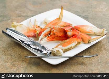Horizontal photo of freshly cooked Dungeness crab legs, focus on large claw in center, on white plate with stainless crab crackers and stone counter top underneath