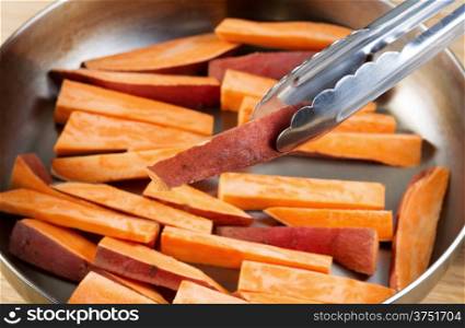 Horizontal photo of freshly cut Yams in stainless steel frying pan with focus on single piece being place into pan with tong