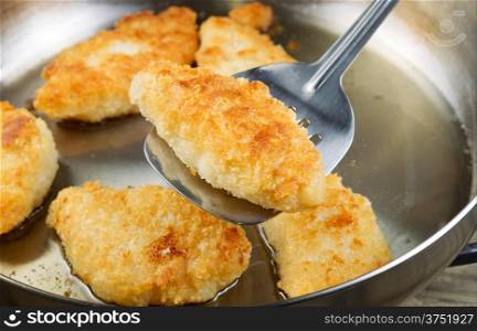 Horizontal photo of golden breaded coated fish being fried in stainless steel frying pan with focus on single piece with spatula underneath