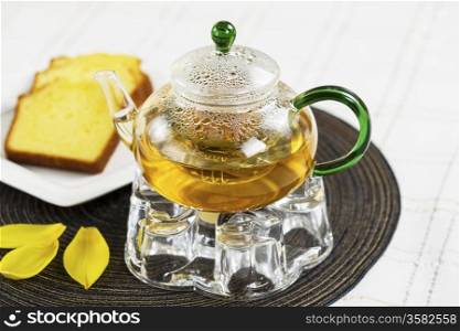 Horizontal photo of Green Tea pot (focus on tea pot), Golden Lemon Pound Cake, yellow leafs on place mat with White Striped Table Cloth in background