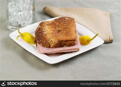 Horizontal photo of ham sandwich made with whole grain bread on white plate with yellow peppers and glass of water in background