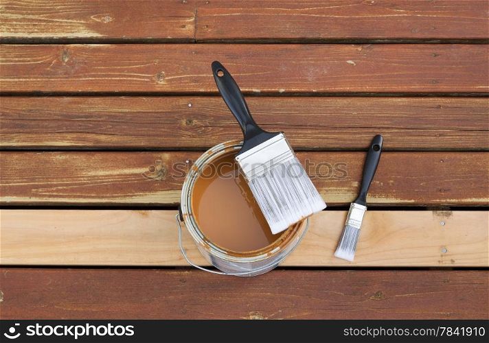 Horizontal photo of large nylon paint brush on top of wood stain can with small paint brush lying on a single new cedar wooden board next to fading wood on outdoor deck