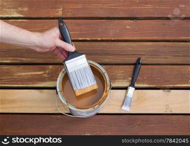 Horizontal photo of male hand dipping paint brush into a can of wood stain with single small brush lying on a single new cedar wooden board next to fading wood on outdoor deck