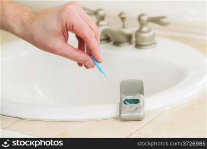 Horizontal photo of male hand picking up dental tooth pick in bathroom with dental floss container, sink and counter top in background