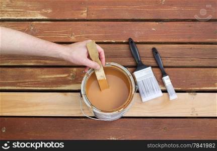 Horizontal photo of male hand stirring wood stain in can with stir stick with two paint brushes lying on a single new cedar wooden board next to fading wood on outdoor deck