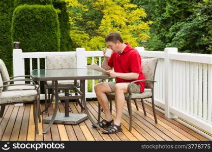 Horizontal photo of mature man drinking morning coffee while reading a magazine on outdoor patio with green and yellow trees in full seasonal bloom