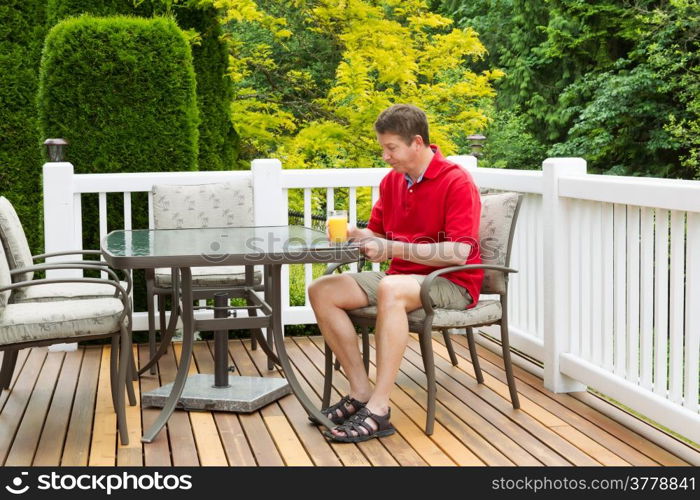 Horizontal photo of mature man holding a glass of orange juice while preparing to read a magazine on outdoor patio with green and yellow trees in full seasonal bloom