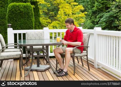 Horizontal photo of mature man holding a glass of orange juice while preparing to read a magazine on outdoor patio with green and yellow trees in full seasonal bloom