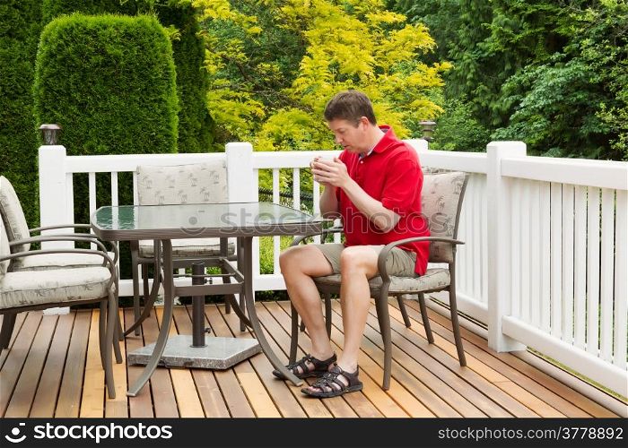 Horizontal photo of mature man holding coffee of hot coffee while preparing to read a magazine on outdoor patio with green and yellow trees in full seasonal bloom