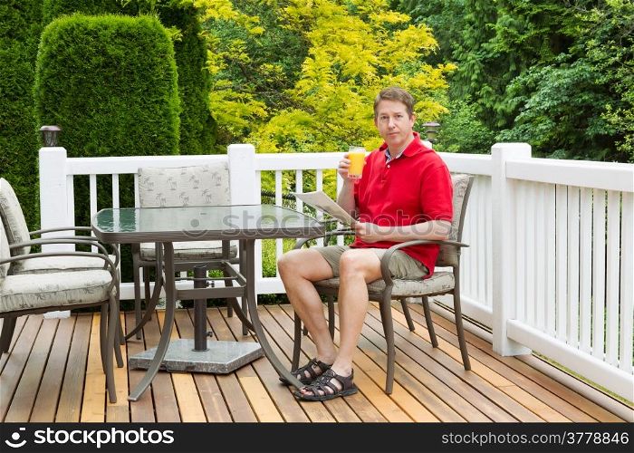 Horizontal photo of mature man, looking forward, while holding a glass of orange juice and magazine on outdoor patio with green and yellow trees in full seasonal bloom