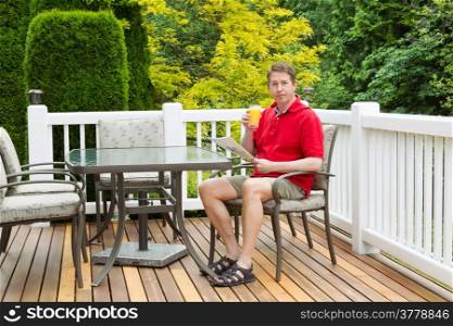 Horizontal photo of mature man, looking forward, while holding a glass of orange juice and magazine on outdoor patio with green and yellow trees in full seasonal bloom