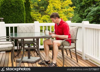 Horizontal photo of mature man reading magazine with cup of coffee in hand on outdoor patio with green and yellow trees in full seasonal bloom
