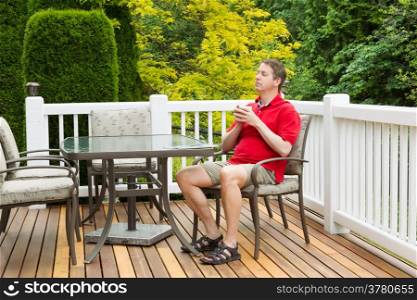 Horizontal photo of mature man, resting in chair, while holding a cup of coffee in his hands on outdoor patio with green and yellow trees in full seasonal bloom