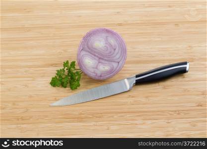 Horizontal photo of sliced purple onion and small piece of parsley with single knife lying on natural bamboo cutting board