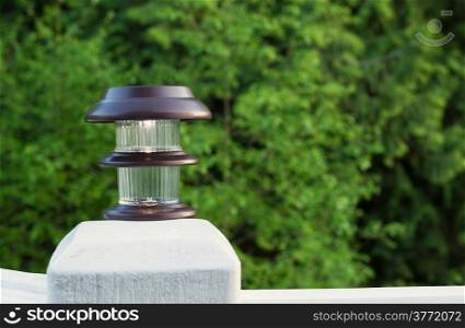 Horizontal photo of solar lamp, light just turning on, on patio post with green trees in background