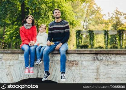 Horizontal portrait of adorable child sits between her mother and father, enjoy beaitiful landscapes from top of ancient bridge, breath fresh air, look into distance. Family and relationship concept