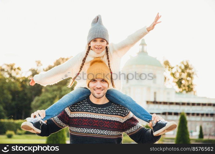 Horizontal portrait of affectionate father gives piggy back ride to his daughter outside, play together, have fun. Happy little charming girl spends weekends with father, waves excitedly with hand