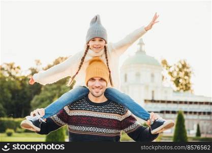 Horizontal portrait of affectionate father gives piggy back ride to his daughter outside, play together, have fun. Happy little charming girl spends weekends with father, waves excitedly with hand