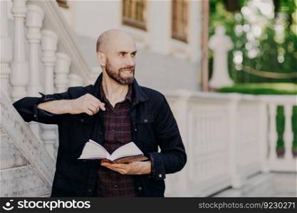 Horizontal portrait of attractive cheerful bald bearded male holds book, looks happily aside, enjoys calm atmosphere in royal park, reads something interesting, concentrated away. Reading concept