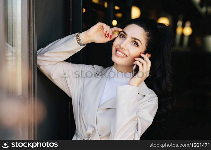 Horizontal portrait of beautiful lady with dark hair and eyes, make-up and lips smiling broadly holding hand on head while speaking over smartphone with her husband. Pretty woman in elegant clothes