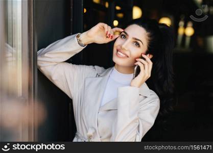 Horizontal portrait of beautiful lady with dark hair and eyes, make-up and lips smiling broadly holding hand on head while speaking over smartphone with her husband. Pretty woman in elegant clothes