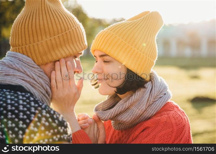 Horizontal portrait of brunette female in warm hat, scarf and sweater, keeps hands on boyfriend`s cheek, have romantic moment, going to kiss as dating. Couple in love look at each other passionately