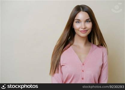 Horizontal portrait of charming European woman smiles gently at camera, has long straight hair, wears casual rosy sweater, going to have outdoor walk, stands against beige studio background.