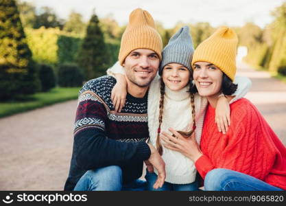 Horizontal portrait of friendly affectionate family hug each other, wear knitted caps and sweaters, walk together, have pleasant smiles on faces. Married couple and small daughter enjoy free time