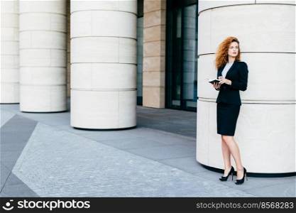 Horizontal portrait of good-looking businesswoman dressed in formal clothes and black shoes with high heels, holding pocket book, looking aside with thoughtful expression. Career and business concept