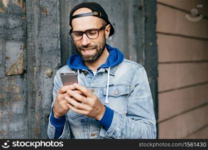 Horizontal portrait of handsome bearded stylish man in denim anorak wearing big eyeglasses holding smartphone using free Internet, checking e-mail, watching funny videos, updating applications