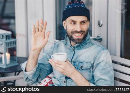 Horizontal portrait of happy male with dark beard and mustache, waves with hand, glad to notice friend, holds cup of tea, wears fashionable clothing. Man greets someone. Hipster guy drinks coffee