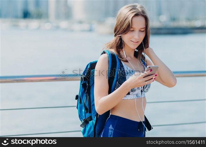 Horizontal portrait of lovely female with perfect figure, wears leggings and top, has focused look into smart phone, carries big rucksack, tries to find correct route while covers long distance alone