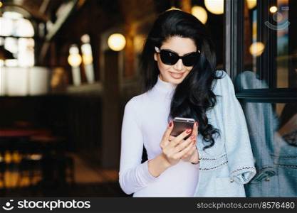 Horizontal portrait of magnificent brunette woman in white clothes and sunglasses sitting in cafe using her smartphone messaging with friends using free internet conncection. Beauty and youth concept