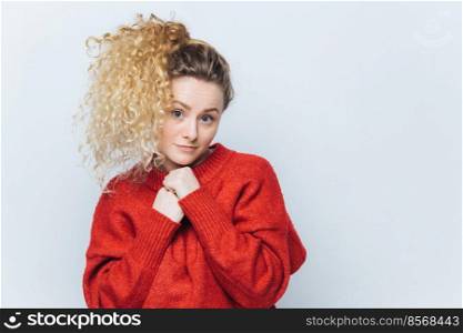 Horizontal portrait of pleasant looking young female with curly light pony tail, dressed in red sweater, looks with non confident expression at camera, poses against white wall with blank copy space