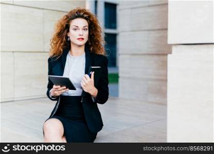 Horizontal portrait of pretty confident businesswoman dressed formally, holding her pocket book with pen and tablet computer, being busy with her work. People, lifestyle, career, business concept