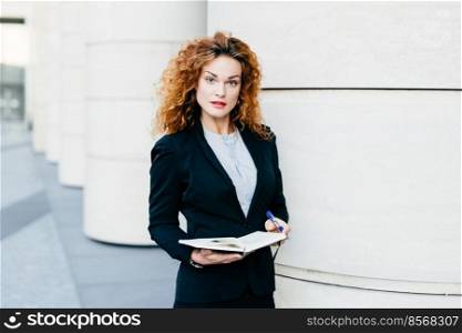 Horizontal portrait of serious pretty businesswoman with curly hair, thin eyebrows and curly hair, wearing black suit and white blouse, holding notebook with pen in hands, looking directly into camera