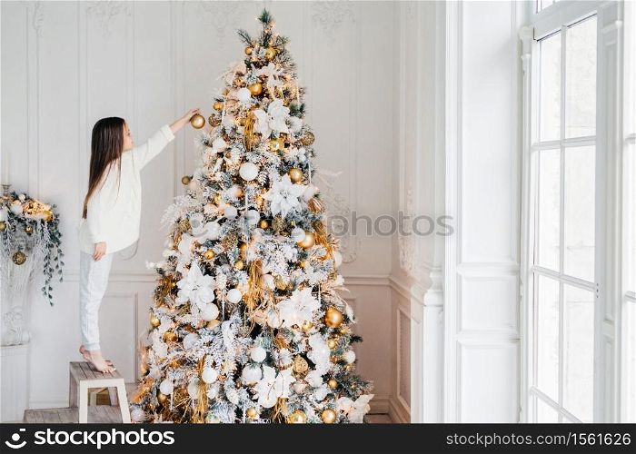 Horizontal portrait of small female child stands on chair, decorates Christmas tree, tries to show best, being at home, enjoys domestic calm atmosphere, wants to satisfy parents, being hard working