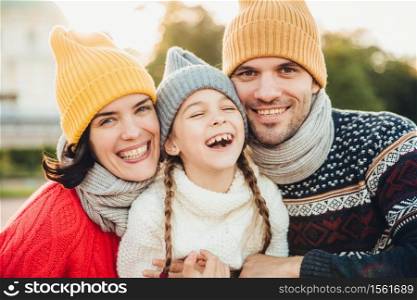 Horizontal portrait of smiling happy woman and man have good time with their daughter, wear knitted hats, warm sweaters and scarfs, going in park to have walk together. Spending time in family circle