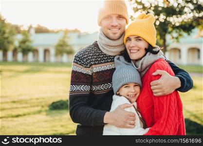 Horizontal portrait of young handsome man in yellow hat and warm knitted sweater, embrace his wife and daughter, pose at camera while stand in park. Three family members spend weekends together