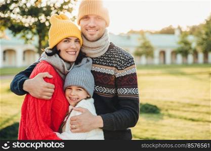 Horizontal portrait of young handsome man in yellow hat and warm knitted sweater, embrace his wife and daughter, pose at camera while stand in park. Three family members spend weekends together